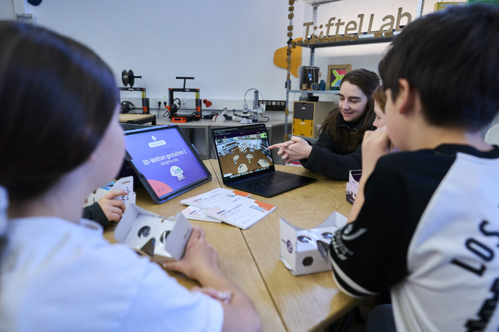 Eine Lehrkraft Sitzt An Einem Gruppentisch Mit 2 Kindern Und Zeigt Auf Einen Laptop. Sie Erklärt Das Programm, Das Auf Dem Laptop Läuft.